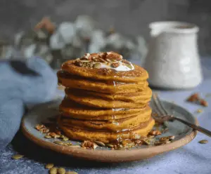Fluffy vegan pumpkin pancakes topped with maple syrup and fresh cinnamon, served with sliced bananas and walnuts on a rustic plate.
